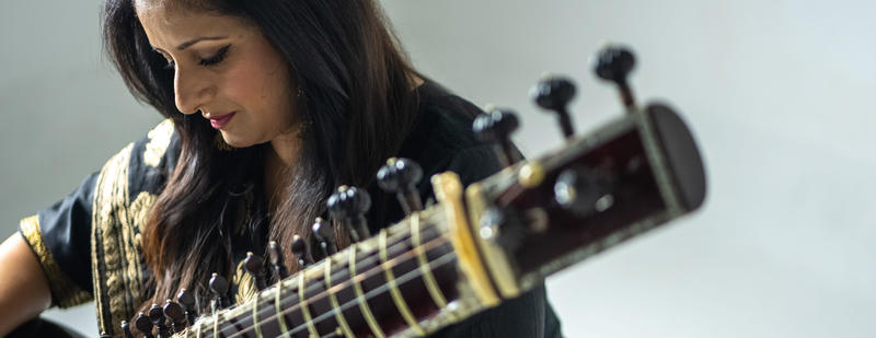A woman dressed in traditional Indian clothing looking down towards her hands, she holds a sitar partially out of frame with the neck and head close to the camera slightly blurred