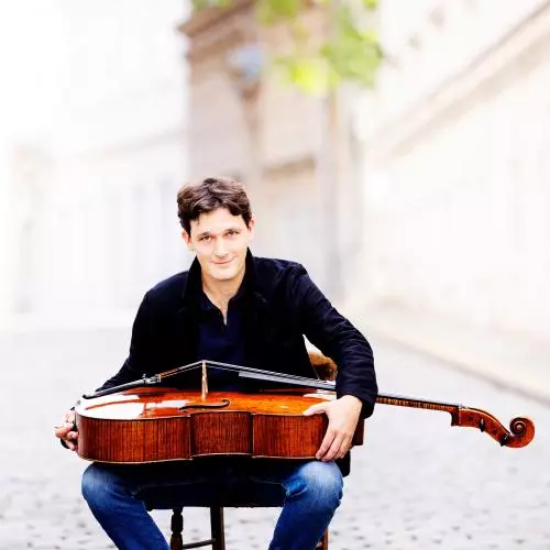 Man in black shirt and jeans sitting on a chair in the middle of a cobbled street holding a cello on his lap